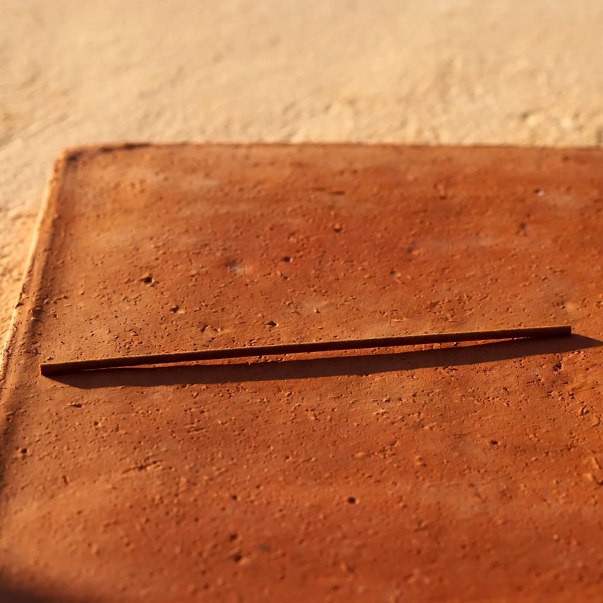 A single long incense stick lying across a terracotta tile